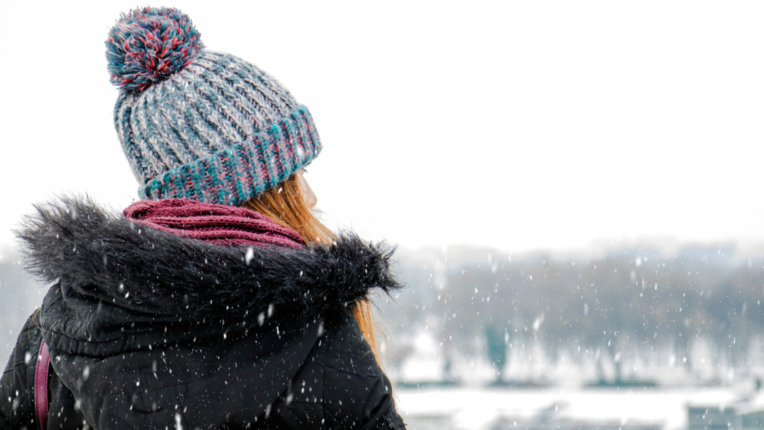 A female presenting person wearing winter clothing looks over a snowy landscape